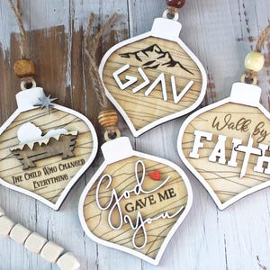 May include: Wooden Christmas ornaments with white borders and various designs. One features a nativity scene with the text "The Child Who Changed Everything." Another says "G>AV" with a mountain graphic. Others say "God Gave Me You" and "Walk by Faith."
