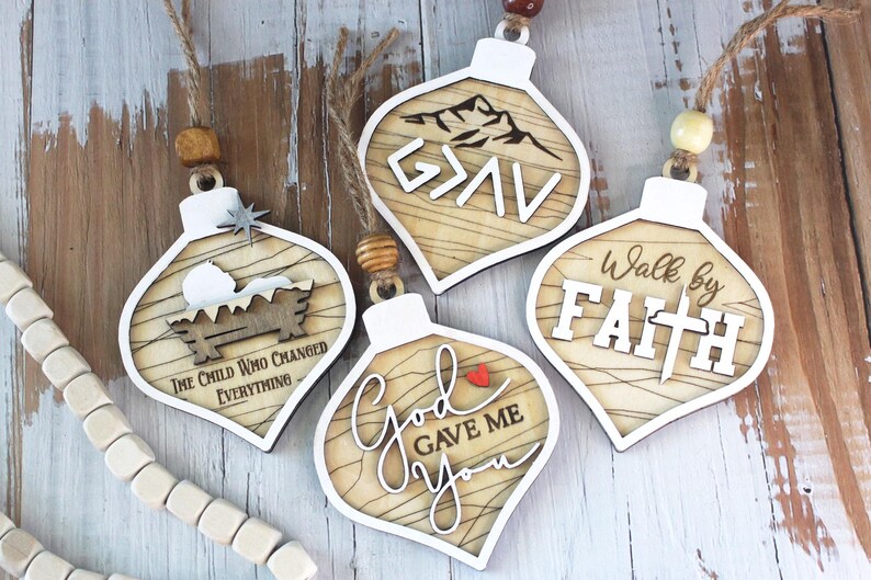 May include: Wooden Christmas ornaments with white borders and various designs. One features a nativity scene with the text "The Child Who Changed Everything." Another says "G>AV" with a mountain graphic. Others say "God Gave Me You" and "Walk by Faith."