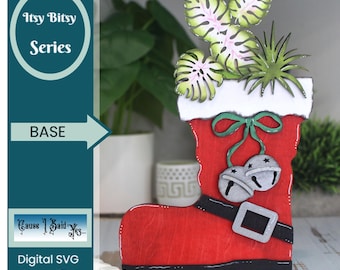 May include: A red wooden Santa boot planter with a white cuff, black belt, and silver jingle bells. The boot holds paper cut-out plants with green and pink leaves. The text "Itsy Bitsy Series" and "Digital SVG Laser File" are visible.
