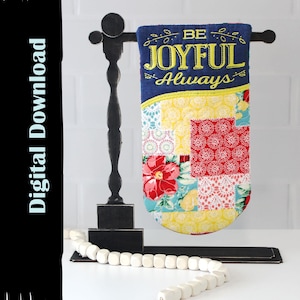 Pode incluir: Uma bandeira decorativa com o texto "BE JOYFUL Always" bordado em amarelo. A bandeira tem um design de patchwork com tecido floral e estampado. Ela está pendurada em um suporte de madeira preta com uma guirlanda de contas brancas.