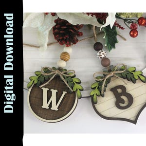 May include: Wooden Christmas ornament with the letter "W" and "B". The ornaments are brown and cream-colored, with green leaf accents and twine bows. The ornaments are strung together with wooden beads and decorative foliage.