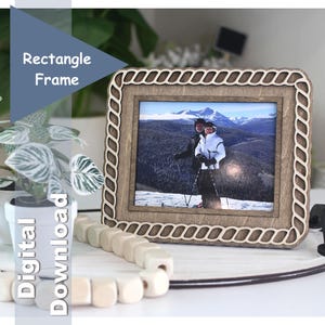 May include: A rectangular wooden photo frame with a decorative rope-like border. The frame holds a photograph of two people skiing in the mountains. The frame is on a white surface with a plant and wooden beads.
