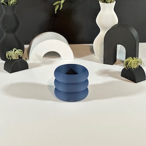 May include: An artistic display of modern home decor items. The arrangement includes black and white vases with greenery, a blue stacked ring sculpture, and geometric shapes. The items are set against a white surface and a dark gray backdrop.