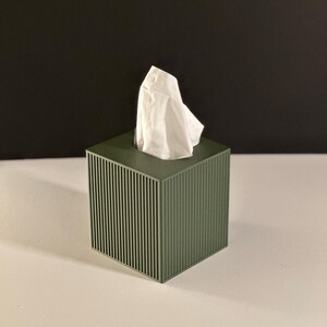 K&ouml;nnte beinhalten: Ein gr&uuml;ner Taschentuchspender mit vertikalem Streifendesign. Wei&szlig;e Taschent&uuml;cher ragen aus der oberen &Ouml;ffnung heraus. Die Box ist quadratisch und steht auf einer wei&szlig;en Oberfl&auml;che mit schwarzem Hintergrund.