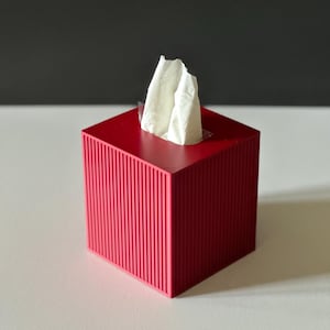 K&ouml;nnte beinhalten: Ein roter Taschentuchspender mit gerilltem Au&szlig;endesign. Wei&szlig;e Taschent&uuml;cher ragen aus der oberen &Ouml;ffnung heraus. Die Box ist quadratisch und steht auf einer wei&szlig;en Oberfl&auml;che mit dunklem grauem Hintergrund.