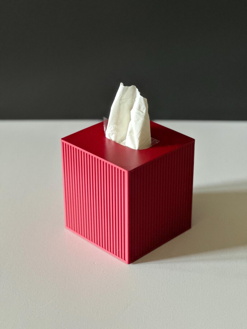 K&ouml;nnte beinhalten: Ein roter Taschentuchspender mit gerilltem Au&szlig;endesign. Wei&szlig;e Taschent&uuml;cher ragen aus der oberen &Ouml;ffnung heraus. Die Box ist quadratisch und steht auf einer wei&szlig;en Oberfl&auml;che mit dunklem grauem Hintergrund.