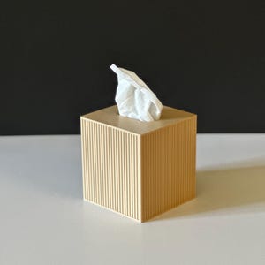 K&ouml;nnte beinhalten: Beige quadratische Taschentuchbox mit vertikalen Rillen. Ein wei&szlig;es Taschentuch ragt aus der Oberseite heraus. Die Box steht auf einer wei&szlig;en Oberfl&auml;che mit schwarzem Hintergrund.