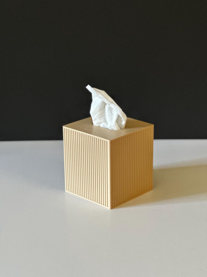 K&ouml;nnte beinhalten: Beige quadratische Taschentuchbox mit vertikalen Rillen. Ein wei&szlig;es Taschentuch ragt aus der Oberseite heraus. Die Box steht auf einer wei&szlig;en Oberfl&auml;che mit schwarzem Hintergrund.