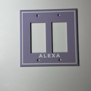 May include: A light purple double light switch wall plate featuring a white rectangular border and the word ALEXA printed in white capital letters below the two rectangular switch openings. The plate has four screw holes for mounting purposes.