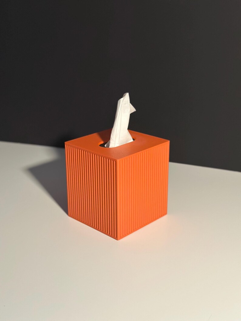 K&ouml;nnte beinhalten: Eine orangefarbene Taschentuchbox mit gerilltem Design. Ein wei&szlig;es Taschentuch ragt aus der oberen &Ouml;ffnung heraus. Die quadratische Box steht auf einer wei&szlig;en Oberfl&auml;che mit schwarzem Hintergrund.