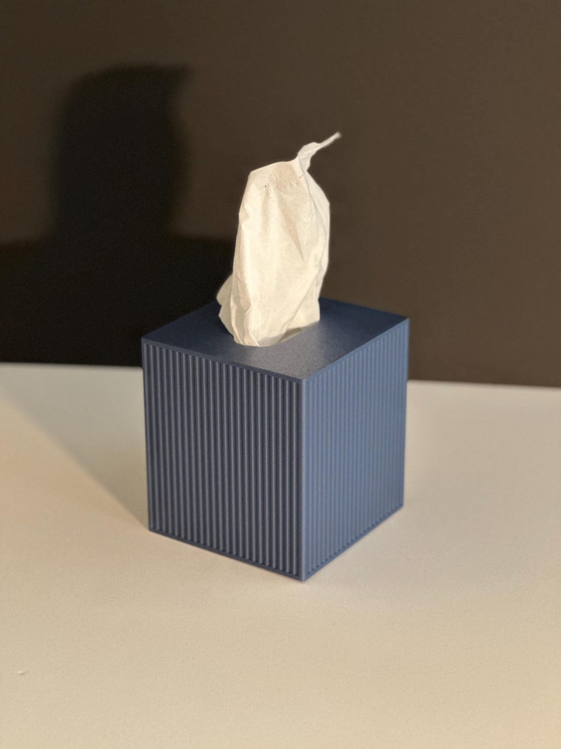 K&ouml;nnte beinhalten: Ein blauer Taschentuchspender mit geripptem Design. Wei&szlig;e Taschent&uuml;cher ragen aus der Oberseite heraus. Die Box ist quadratisch und steht auf einer wei&szlig;en Oberfl&auml;che.