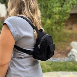 May include: A black corduroy mini backpack with a zipper closure and a flap pocket. The backpack is worn by a person with long blonde hair.