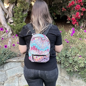 May include: A small backpack with a colorful geometric pattern in shades of blue, pink, and white. The backpack has black straps and a small front pocket with a black zipper. The person is wearing a black shirt and dark jeans.