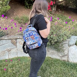 May include: A small backpack with a blue, white, and orange tile pattern. The backpack has a black strap and a black zipper. The person is wearing a black shirt and dark jeans. The background includes a stone wall, green grass, and flowers.