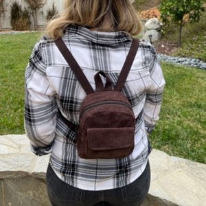 May include: A brown corduroy backpack with adjustable straps and a front pocket. The backpack is worn over a black and white plaid shirt. The bag has a zippered closure and a top handle.