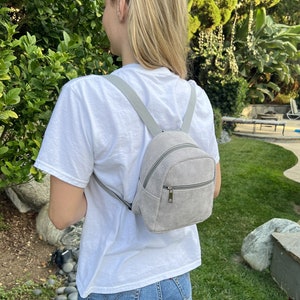 May include: A gray corduroy mini backpack with a zipper closure. The backpack has a small, rectangular shape and is worn by a person.
