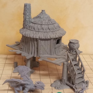 Könnte beinhalten: Eine graue 3D-gedruckte Miniatur einer Strohdachhütte auf Stelzen mit einem Kessel und einer Treppe. Im Vordergrund befindet sich eine graue Drachenminiatur mit fünf Würfeln davor.