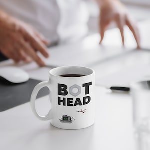 May include: White ceramic mug with the text "BOT HEAD" in black block letters, a silver nut graphic, and small robot and drone illustrations. The mug is filled with dark liquid and sits on a white desk.