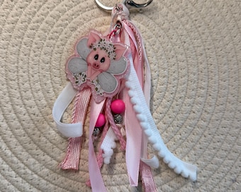 Peut inclure: Porte-clés décoratif rose et blanc avec un motif de cochon. Le cochon est dans un cadre en forme de fleur avec des détails en paillettes argentées. Le porte-clés comprend des rubans roses et blancs, des pompons et des perles roses, attachés à un porte-clés argenté.