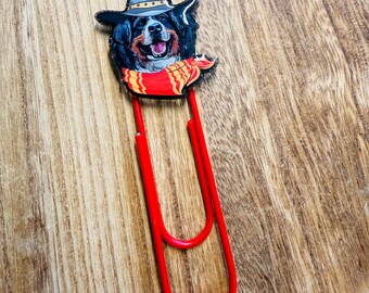 Peut inclure: Une attache-papier rouge en métal représentant un chien souriant portant un chapeau de cow-boy et une écharpe rouge avec des détails jaunes. Le chien a des poils noirs et bruns. L'attache-papier est sur une surface en bois.