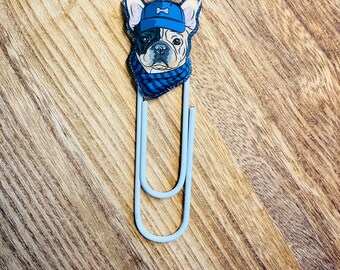 Peut inclure: Un trombone bleu clair avec un motif de bouledogue. Le bouledogue porte une casquette bleue et une écharpe à carreaux bleus et noirs. Le visage du bouledogue est noir, blanc et beige. Le trombone est posé sur une surface en bois.