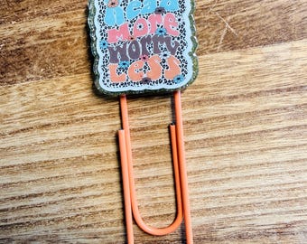 Peut inclure: Un trombone orange avec un charme rectangulaire. Le charme porte les mots "read more worry less" en bleu, rose et orange, avec des motifs floraux et un fond léopard. Un accessoire de papeterie.