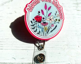 Peut inclure: Un enrouleur de badge rond rose et blanc avec l'inscription "bloom with kindness" en rose. Le centre présente un motif floral avec des fleurs rouges, roses et jaunes sur un fond bleu clair. Comprend un clip métallique et une sangle en plastique transparent.
