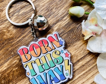 Peut inclure: Un porte-clés arc-en-ciel avec les mots "Born This Way" dans une police rétro. Le porte-clés est recouvert de paillettes et d'étoiles et est attaché à un anneau de clé argenté avec un charme de boule disco.