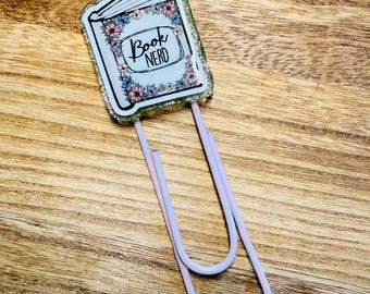 Peut inclure: Un trombone décoratif avec un motif de livre et les mots "Book Nerd" avec des accents floraux. Le clip a une bordure scintillante et une base de trombone rose clair. Le fond est une surface en bois.