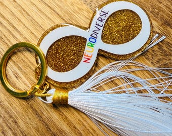 Peut inclure: Un porte-clés symbole infini doré à paillettes avec le mot "NEURODIVERSE" en couleurs arc-en-ciel. Le porte-clés a une bordure blanche et un pompon blanc avec un accent doré. L'anneau porte-clés est doré.