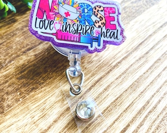Peut inclure: Un porte-badge violet et scintillant avec le mot "NURSE" en rose, bleu et imprimé léopard. Le texte "Love inspire heal" est également inclus. Le porte-badge présente une seringue, un chapeau d'infirmière et un accent de strass.