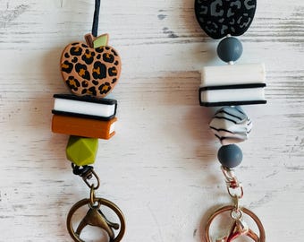 Peut inclure: Deux porte-clés à perles avec des breloques en forme de pomme et de livre. L'un présente une pomme à imprimé léopard, des livres orange et blancs et un fermoir de couleur bronze. L'autre a une pomme noire et grise, des perles blanches et noires et un fermoir en forme de signe de paix.