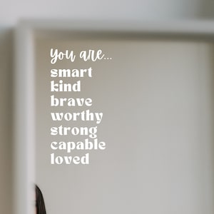 May include: White text on a white wall reads "You are... smart kind brave worthy strong capable loved."