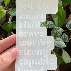 Puede incluir: Pegatina blanca con el texto "You are... smart kind brave worthy strong capable loved" sobre un fondo gris claro.