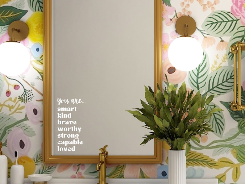 Puede incluir: Un espejo con marco dorado con un fondo de papel tapiz floral. El espejo tiene una calcoman&iacute;a de vinilo blanco con el texto "You are... smart kind brave worthy strong capable loved".