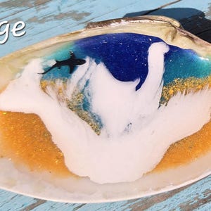 May include: A large, decorative seashell with a painted ocean scene. The interior features a blue and turquoise ocean with a white wave and a small black shark. The base is a golden color, and the word "Large" is written in white.