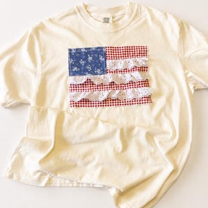 May include: Cream-coloured t-shirt with a fabric American flag design. The flag features a blue floral print square, red and white gingham, and white eyelet lace stripes. The shirt is made of soft material.