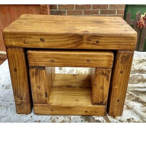 May include: A set of two rustic wooden nesting tables with a natural wood finish. The top table is larger and the bottom table is smaller and has an open shelf.