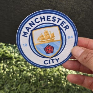May include: A round patch featuring the Manchester City Football Club logo. The logo includes a shield with a ship, a red rose, and the words "Manchester City" in a blue and white circular border. The numbers "18" and "94" are also visible.