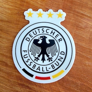May include: A white and black circular sticker with the text "DEUTSCHER FUSSBALL-BUND" and a black eagle inside. There are four gold stars above the circle.