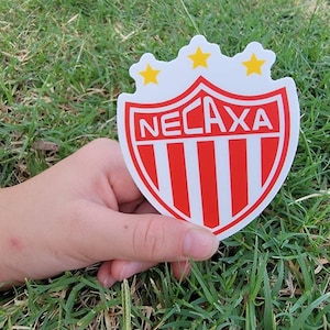 May include: A white and red soccer team logo with the word "NECAXA" in white letters. Three yellow stars are above the word. The logo is on a white background.