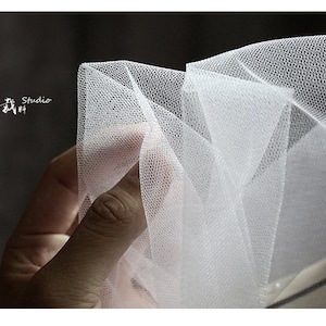 May include: White tulle fabric with a fine mesh texture. The fabric is folded and draped, creating a soft and delicate look.