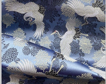 May include: Blue fabric with a white and silver brocade pattern of cranes and flowers. The fabric is folded and has a soft, silky texture.
