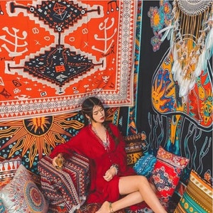 May include: A woman in a red dress sits on a colorful patterned rug with pillows. The rug and pillows feature a variety of geometric and floral designs. The background is decorated with tapestries featuring similar patterns.
