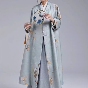 May include: A long, light blue coat with a floral print. The coat has a mandarin collar and a button closure. The coat is worn over a gray skirt.