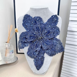 May include: A large, navy blue floral applique, heavily embellished with sequins and beads, is displayed on a gray mannequin bust. The flower has multiple layered petals and a detailed center, creating a textured and dimensional effect. The design is likely for fashion or craft projects.