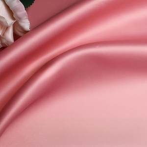 May include: A smooth, lustrous fabric in a solid dusty rose pink colour with deep, flowing folds and ripples. A single, large, pale pink artificial rose flower with layered petals is positioned in the upper left corner of the frame.
