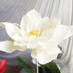 以下が含まれることがあります： 複数の層状の花びらを持つ、大きな白い人工の蓮の花。 花の中心は黄色で、小さな赤い点があります。 花は細い金属の茎にあります。 テキスト「MHONTILY」が左上隅に表示されます。