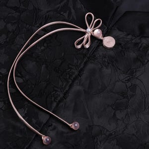 Op de afbeelding: Close-up van zwarte stof met bloemenpatroon. Een lichtroze kikker sluiting met een parel accent en twee bungelende roze kralen is bevestigd aan de stof. De sluiting is gemaakt van een lintachtig materiaal.