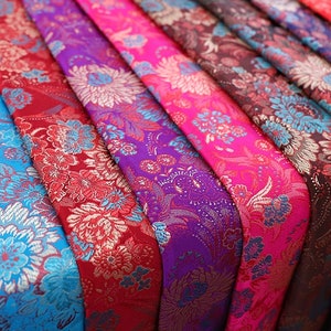 Könnte beinhalten: Nahaufnahme von buntem Stoff mit Blumenmustern. Der Stoff ist fächerförmig angeordnet, mit Farben von Blau über Lila und Rosa bis Rot. Der Stoff hat eine glänzende, satinartige Oberfläche.